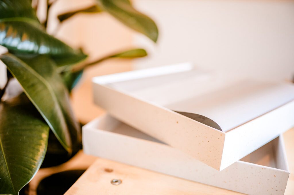 Een minimalistische witte bewaardoos met een trouwalbum erin, liggend naast een groene plant op een houten tafel.