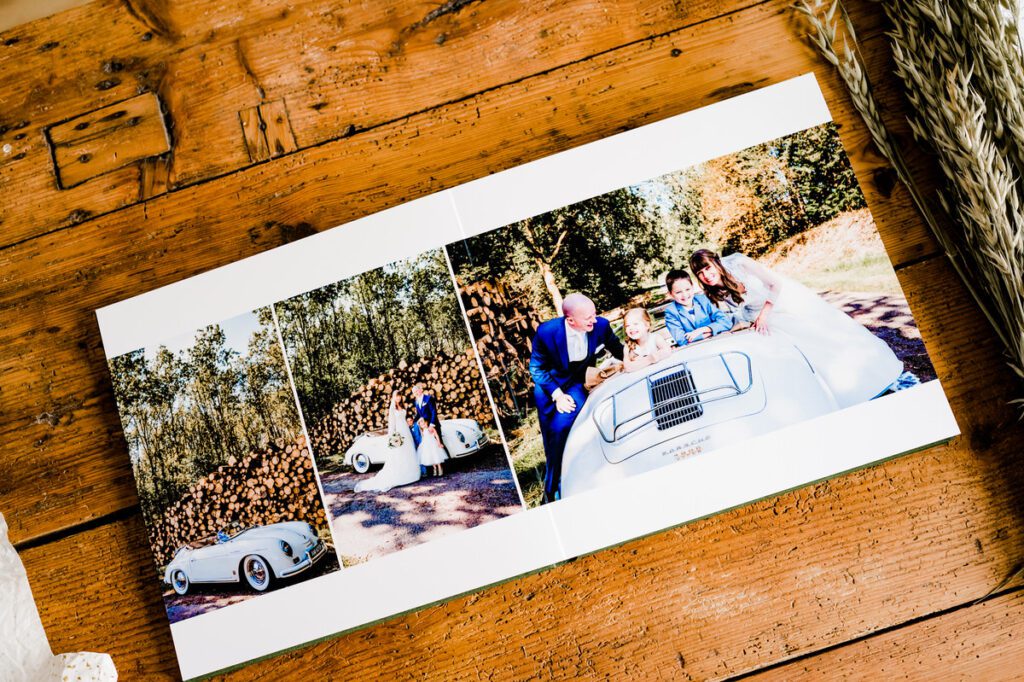 Open trouwalbum met kleurrijke foto's van een bruidspaar en hun kinderen, naast een klassieke cabriolet, liggend op een houten tafel.