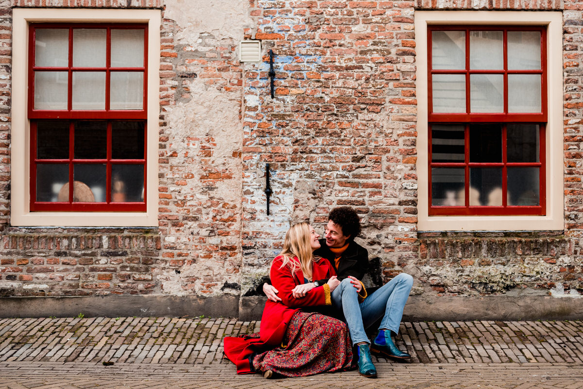 Een stel zit op straat tegen een oude bakstenen muur met rode kozijnen. De vrouw draagt een rode jas en kijkt liefdevol naar de man, die vrolijk terugkijkt.